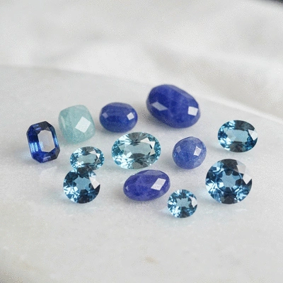 Assortment of polished blue gemstones, including sapphire, aquamarine, tanzanite, and blue topaz, arranged on a smooth, light-colored surface, clean image